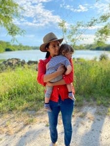 (Image description: Marita standing outside in front of a lake and trees on a sunny day. She is wearing a hat, red shirt, and jeans and is holding her son Reid, who is wearing a blue outfit.)