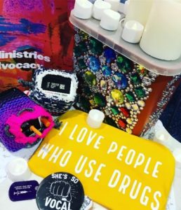 Image description: decorated needle receptacle and naloxone spray, candles, a yellow cloth that says ‘I love people who use drugs’ and other colorful ritual items. 