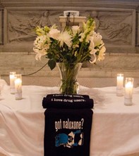 Image description: altar covered in white cloth with vase of flowers, candles, and items which say, ‘got naloxone?’ and ‘I love drug users.’